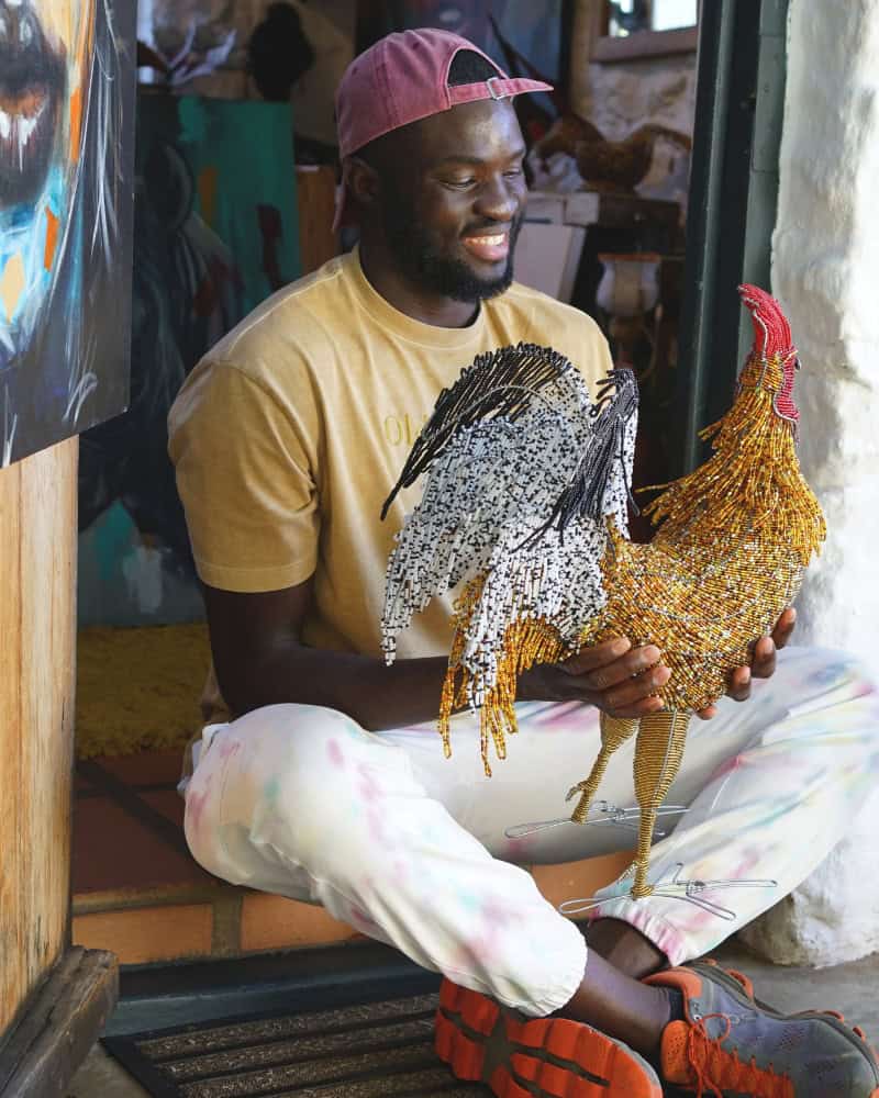 Tinashe Kachama holding a completed hand-beaded rooster sculpture in vibrant reds and oranges